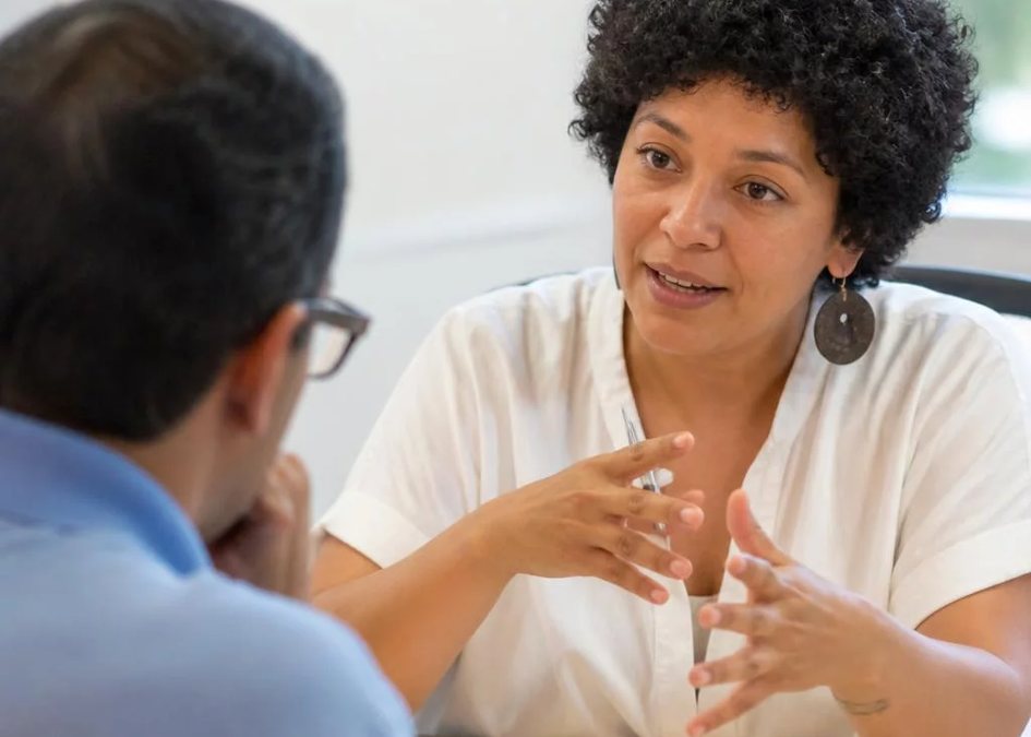 Duas pessoas frente a frente na mesa: uma sustenta a fala, a outra anota o que ouviu